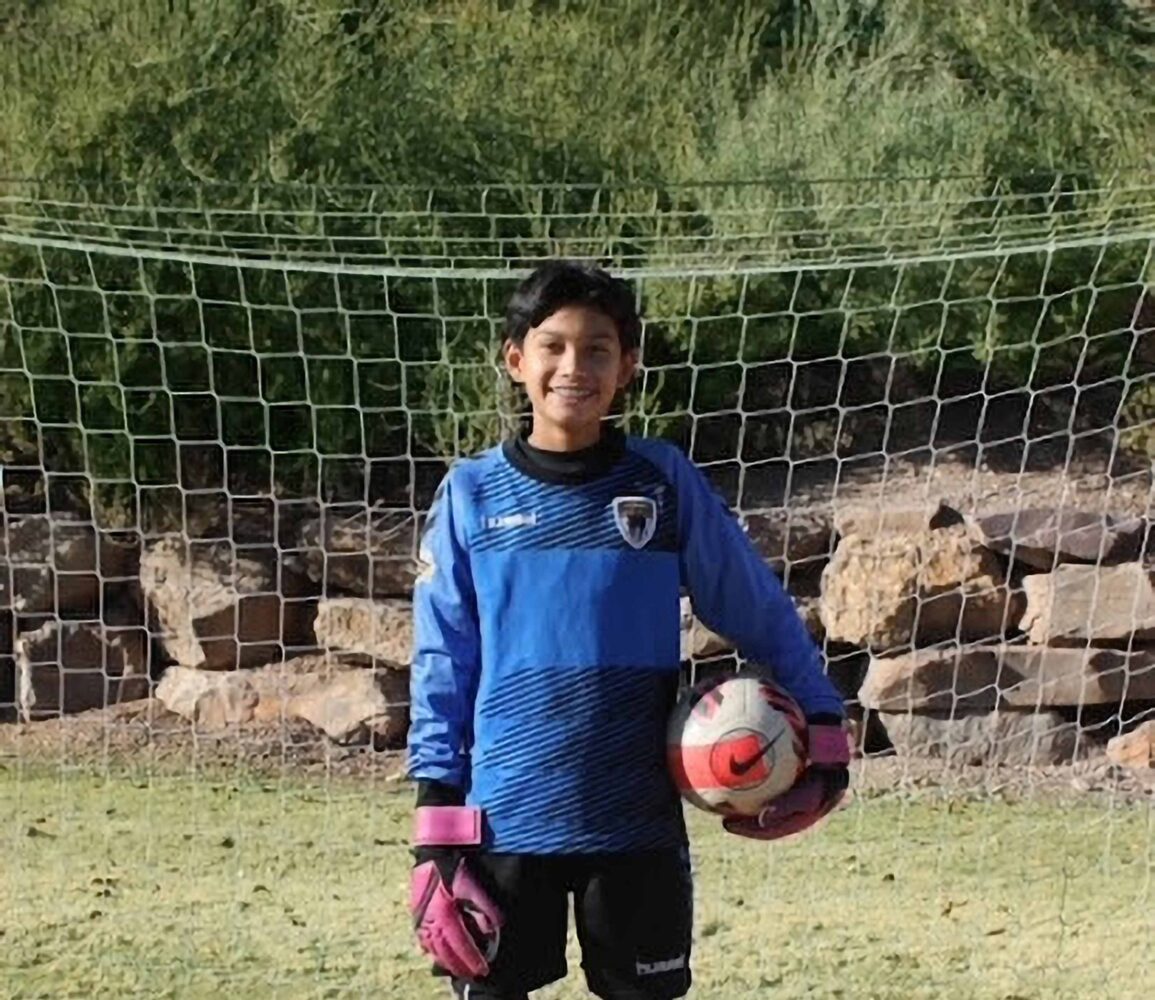 kid goalie holding a ball in his left hand standing under a soccer net